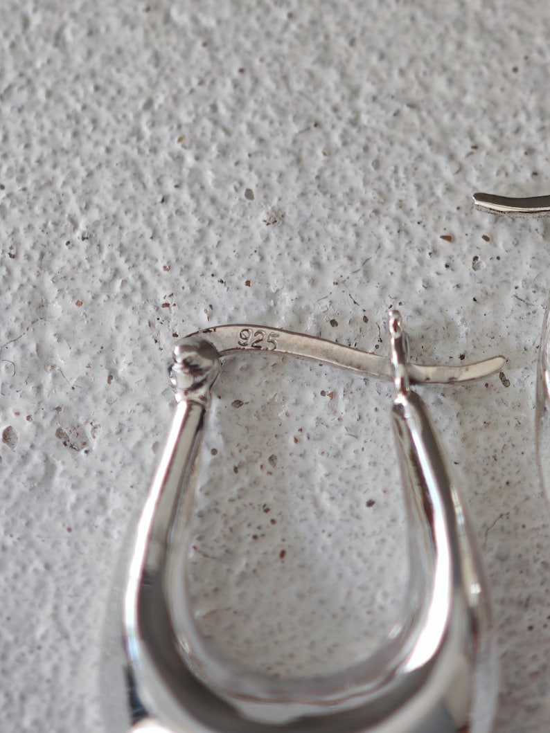 May include: Close-up of a pair of silver hoop earrings. The earrings have a smooth, polished finish and a teardrop shape. The clasp is visible, and the metal is stamped with "925". The background is a textured, off-white surface.