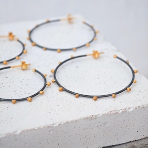 May include: A set of hoop earrings with a dark gray band and small gold-colored beads evenly spaced around the circumference. The earrings are displayed on a white, textured surface.