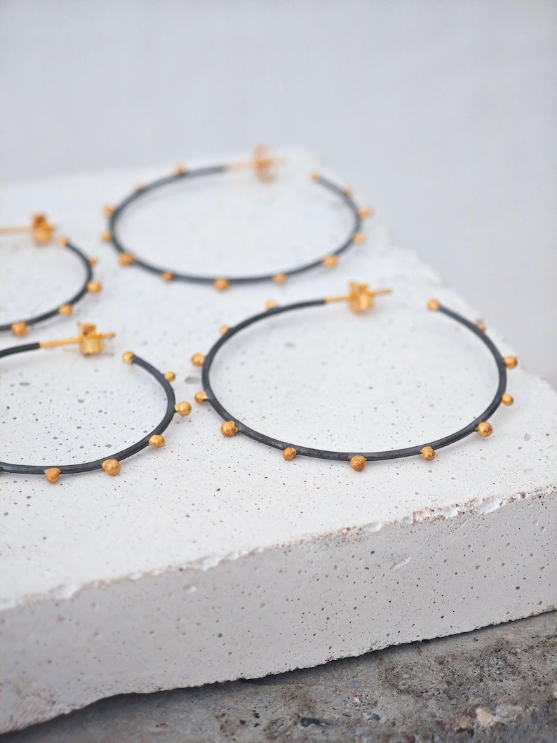 May include: A set of hoop earrings with a dark gray band and small gold-colored beads evenly spaced around the circumference. The earrings are displayed on a white, textured surface.