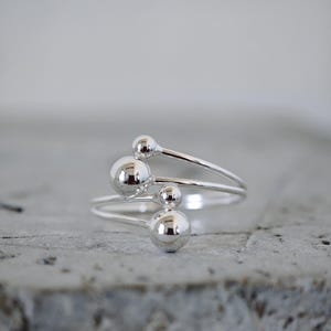 Puede incluir: Un anillo de plata con un diseño moderno. El anillo presenta tres bandas, cada una adornada con un acento esférico brillante. Las esferas varían en tamaño, creando un aspecto único y llamativo. El anillo está sobre un fondo neutro.