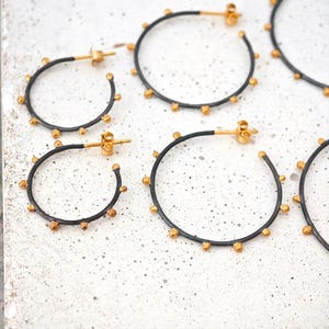 May include: Several pairs of hoop earrings with a black finish and gold-colored accents. The earrings are arranged on a white surface. The earrings have a circular design with small, evenly spaced gold-colored beads around the outer edge.