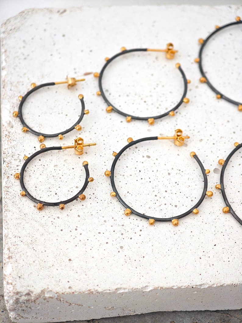 May include: Several pairs of hoop earrings with a black finish and gold-colored accents. The earrings are arranged on a white surface. The earrings have a circular design with small, evenly spaced gold-colored beads around the outer edge.