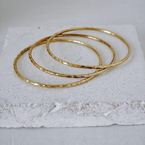 Peut inclure: Trois joncs en métal doré avec une surface martelée et texturée. Chaque bracelet est un anneau circulaire fin avec une finition réfléchissante. Les bracelets sont empilés en se chevauchant légèrement, mettant en valeur l'éclat métallique.