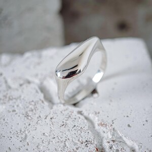Peut inclure: Une bague en argent poli présentant un élément supérieur lisse en forme de goutte d'eau qui se rétrécit en un anneau arrondi. La surface métallique est hautement réfléchissante, affichant une forme propre et continue sans gravures.