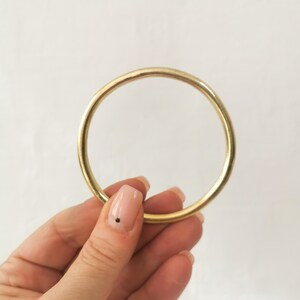 May include: A gold-colored, circular bracelet. The bracelet is a simple, solid band with a smooth surface. The image is set against a plain white background. The bracelet is held by a person.