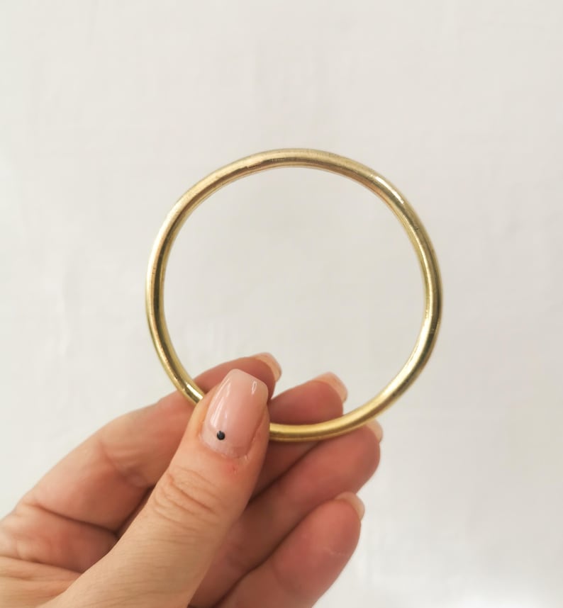 May include: A gold-colored, circular bracelet. The bracelet is a simple, solid band with a smooth surface. The image is set against a plain white background. The bracelet is held by a person.