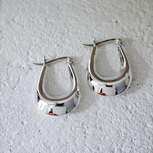May include: A pair of polished silver hoop earrings. The earrings have a teardrop shape with a smooth, reflective surface. The earrings are displayed on a textured, off-white surface.