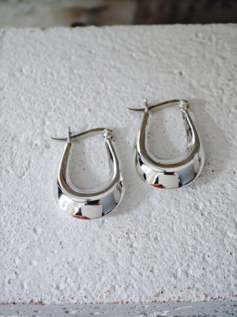 May include: A pair of polished silver hoop earrings. The earrings have a teardrop shape with a smooth, reflective surface. The earrings are displayed on a textured, off-white surface.