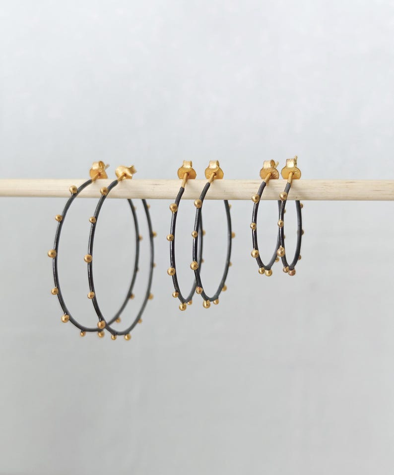 May include: Three pairs of hoop earrings in varying sizes. The earrings have a black metal finish with small, evenly spaced gold-colored beads. The posts and the beads are gold-colored. The earrings are displayed on a wooden dowel against a plain background.