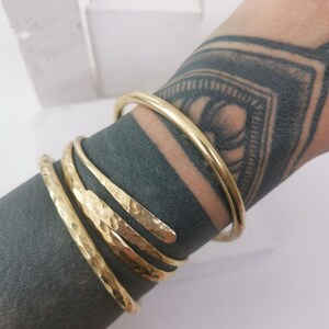May include: A gold-colored bracelet set. The set includes a smooth, round bangle and three hammered-texture, open-ended bracelets. The bracelets are wrapped around a wrist with a dark tattoo.