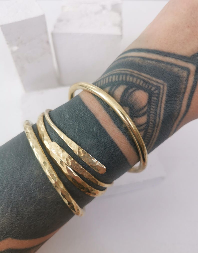 May include: A gold-colored bracelet set. The set includes a smooth, round bangle and three hammered-texture, open-ended bracelets. The bracelets are wrapped around a wrist with a dark tattoo.