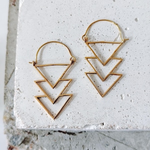 May include: A pair of gold-colored earrings. Each earring features a geometric design with three downward-pointing triangles. The earrings are suspended from a curved hook. The earrings are displayed on a textured, light-colored surface.