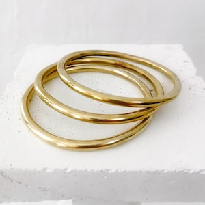 May include: Three gold-colored bangles are stacked on a white surface. The bangles are circular and have a smooth, polished finish. The bangles are a warm, metallic color, and they are arranged in a slightly overlapping manner.