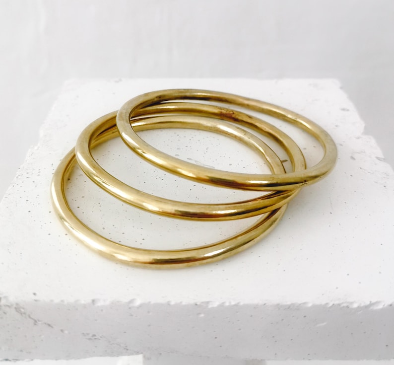 May include: Three gold-colored bangles are stacked on a white surface. The bangles are circular and have a smooth, polished finish. The bangles are a warm, metallic color, and they are arranged in a slightly overlapping manner.