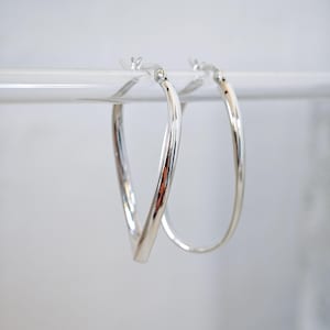 Può includere: Un paio di orecchini a cerchio in tonalità argento lucido con una distinta silhouette a goccia o a forma di V. Gli orecchini presentano una superficie metallica liscia e riflettente con un meccanismo di chiusura a cerniera, appesi a una barra.