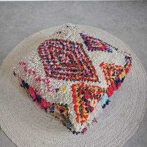 Peut inclure: Un pouf de sol carré doté d'une base épaisse et hirsute blanc cassé avec un motif géométrique multicolore vif dans des tons de rouge, bleu, jaune et rose. La surface texturée présente une construction dense à poils noués sur l'ensemble.