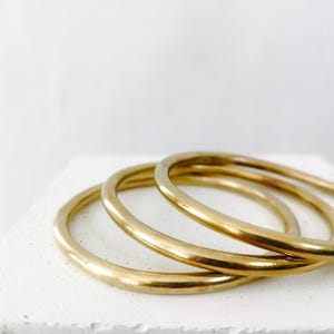 May include: Three gold-colored bangles are stacked on a white surface. The bangles are smooth and circular, reflecting light. The background is a plain white, emphasizing the jewelry's simple elegance.