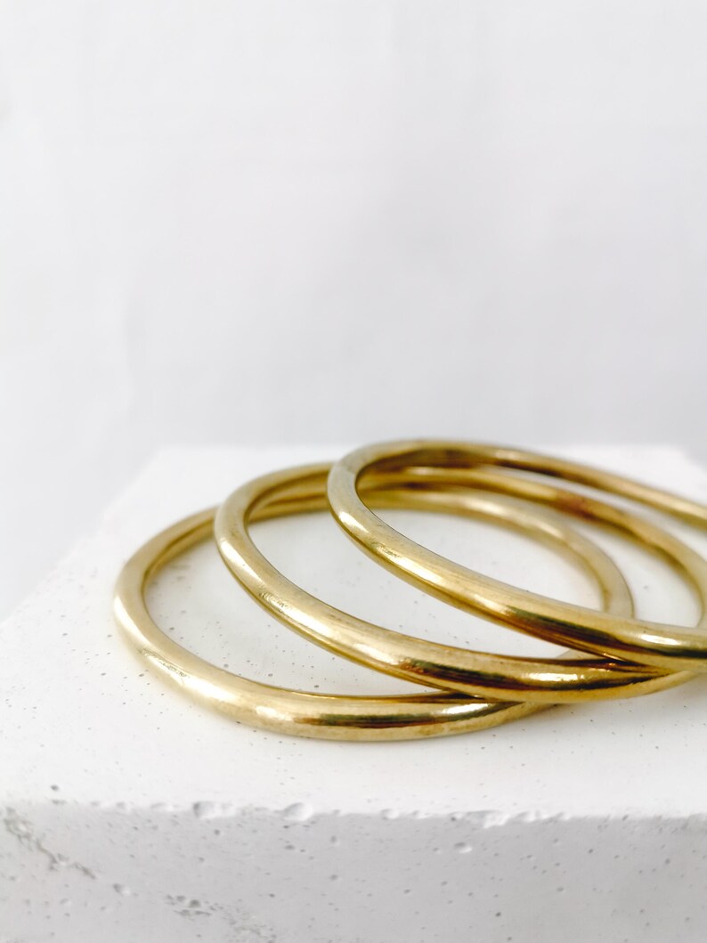 May include: Three gold-colored bangles are stacked on a white surface. The bangles are smooth and circular, reflecting light. The background is a plain white, emphasizing the jewelry's simple elegance.