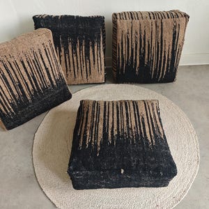 May include: Four square woven ottomans with a black and tan striped design. The ottomans are arranged on a light beige round rug and a gray concrete floor. The ottomans are a neutral color and would fit in any room.
