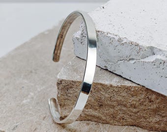 Puede incluir: Un brazalete de plata con una superficie lisa y plana. El brazalete está apoyado en una pila de dos rocas de color marrón claro.