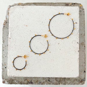 May include: Three sets of hoop earrings with a dark gray finish and small gold-colored accents. The earrings are arranged on a light gray, textured surface. The sizes vary, with the largest hoop measuring approximately 2 inches in diameter.