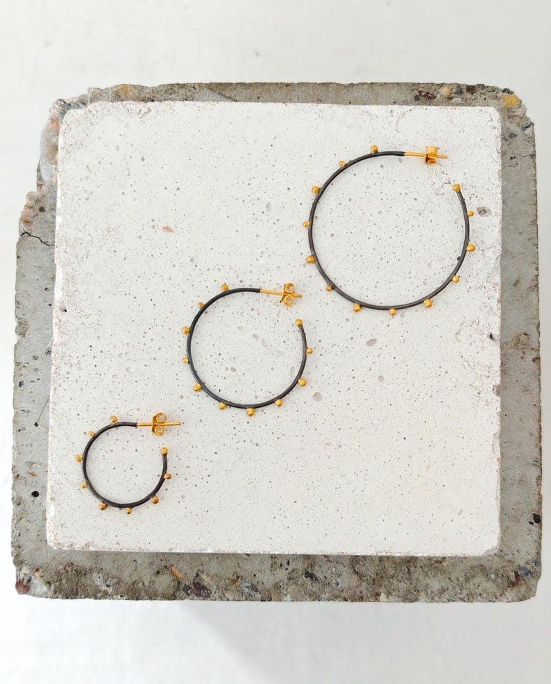 May include: Three sets of hoop earrings with a dark gray finish and small gold-colored accents. The earrings are arranged on a light gray, textured surface. The sizes vary, with the largest hoop measuring approximately 2 inches in diameter.