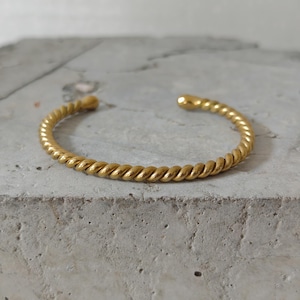 Op de afbeelding: Een goudkleurige manchetarmband met een gedraaid touwontwerp. De open armband is gemaakt van een glanzend, metallic materiaal. De armband ligt op een grijze, getextureerde ondergrond.