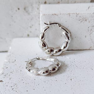 May include: A pair of silver hoop earrings with a textured, beaded design. The earrings are displayed on a white surface, showcasing their shiny, reflective quality. The hoops have a classic, yet modern appeal.