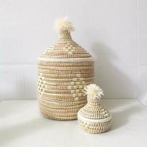 Op de afbeelding: Twee geweven opbergmanden met deksels. De grotere mand is cilindrisch en de kleinere vaasvormig. Beide zijn beige met witte en gouden accenten en afgewerkt met een pluizige, crèmekleurige pompon.