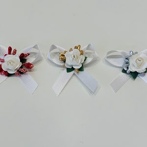 Puede incluir: Tres lazos de satén blanco con una rosa blanca y adornos de diferentes colores. Los lazos están atados con una cinta blanca.