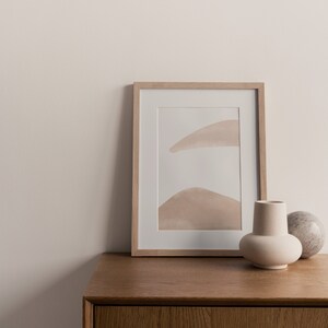 May include: A framed abstract print with a light brown and white color scheme sits on a wooden dresser with a white ceramic vase and a stone object.