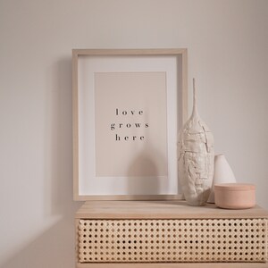 May include: A framed print with the text "love grows here" on a light beige background. The frame is light wood and is sitting on a light wood table with a woven rattan drawer. There are two white ceramic vases and a light pink ceramic box on the table.