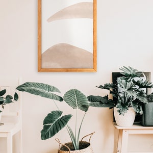 May include: A framed abstract art print with a light brown watercolor design on a white background. The print is hanging on a white wall above a large green plant in a woven basket.