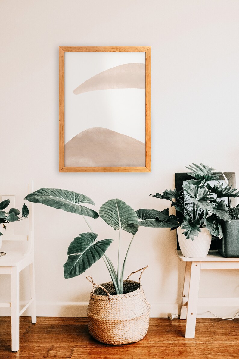 May include: A framed abstract art print with a light brown watercolor design on a white background. The print is hanging on a white wall above a large green plant in a woven basket.