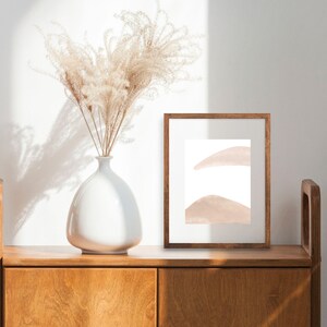 May include: A white ceramic vase with dried pampas grass sits on a wooden cabinet with a framed abstract print. The print features two soft pink shapes on a white background.