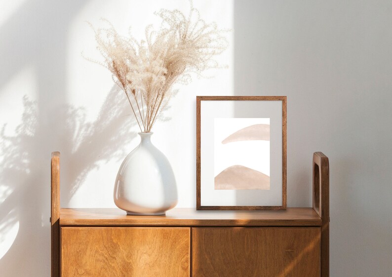 May include: A white ceramic vase with dried pampas grass sits on a wooden cabinet with a framed abstract print. The print features two soft pink shapes on a white background.