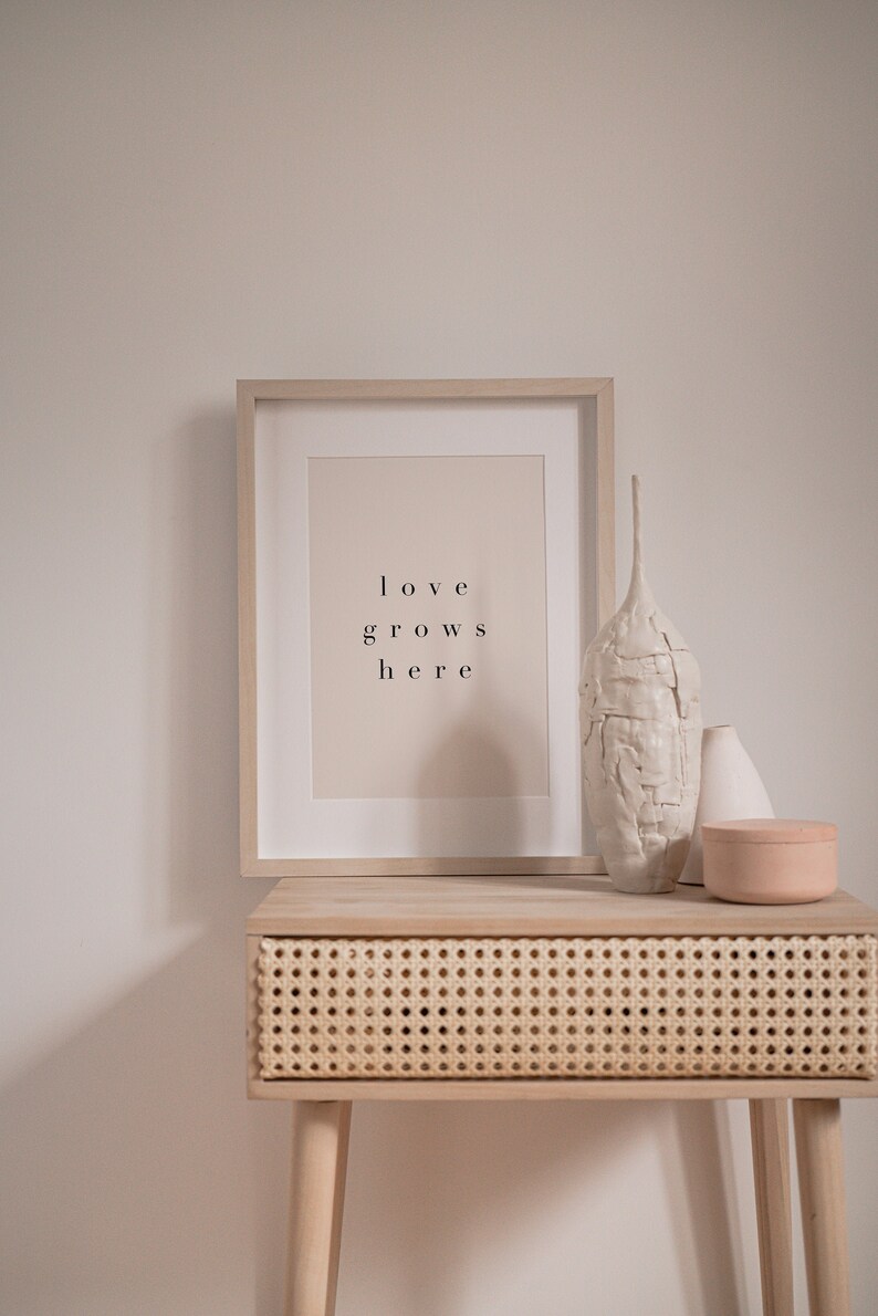 May include: A framed print with the text "love grows here" on a light beige background. The frame is light wood and is sitting on a light wood table with a woven rattan drawer. There are two white ceramic vases and a light pink ceramic box on the table.