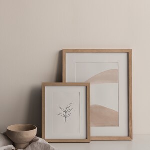 May include: Two framed prints on a white surface. The larger print features a minimalist abstract design with beige and white colors. The smaller print features a simple line drawing of a branch with leaves. A small beige ceramic bowl and dried flowers are in the foreground.