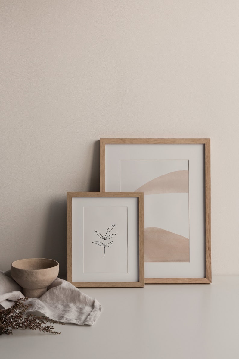 May include: Two framed prints on a white surface. The larger print features a minimalist abstract design with beige and white colors. The smaller print features a simple line drawing of a branch with leaves. A small beige ceramic bowl and dried flowers are in the foreground.