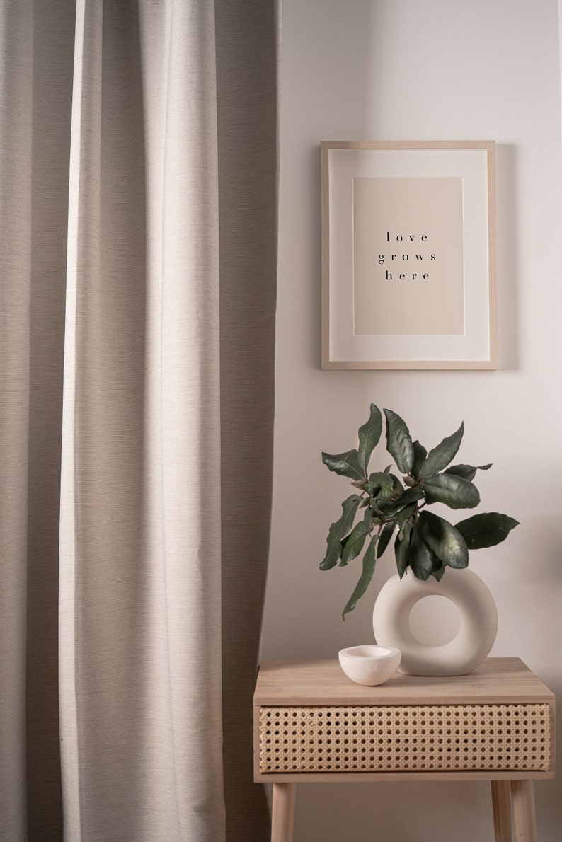 May include: A light brown wooden nightstand with a woven rattan drawer and a white ceramic vase with a ring design. The vase holds a leafy green plant. A framed print with the text "love grows here" hangs on the wall behind the nightstand.