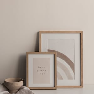 May include: Two wooden picture frames on a white surface. The larger frame contains a watercolor painting of a rainbow in shades of brown. The smaller frame contains a print with the text "love grows here".
