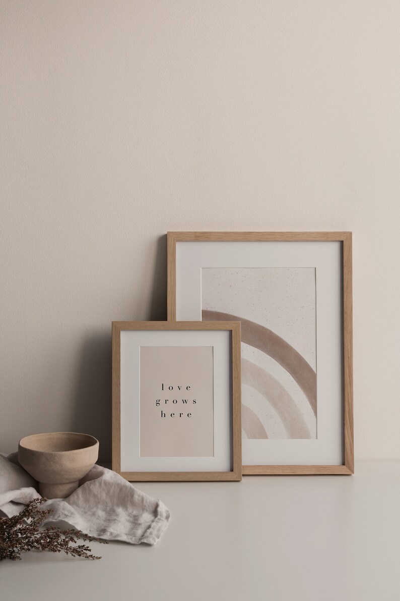 May include: Two wooden picture frames on a white surface. The larger frame contains a watercolor painting of a rainbow in shades of brown. The smaller frame contains a print with the text "love grows here".