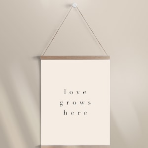 May include: A beige wall with a wooden frame hanging from a string. The frame holds a white poster with the text "love grows here" printed in black.