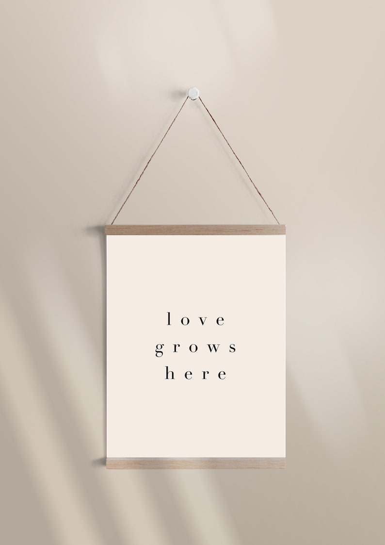May include: A beige wall with a wooden frame hanging from a string. The frame holds a white poster with the text "love grows here" printed in black.
