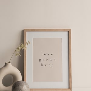 May include: A framed print with the text "love grows here" in a light brown frame on a white surface with a beige vase and a round stone.