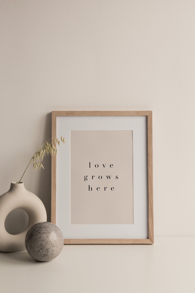 May include: A framed print with the text "love grows here" in a light brown frame on a white surface with a beige vase and a round stone.