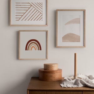 May include: Three framed abstract art prints with a minimalist design. The prints feature geometric shapes and lines in shades of brown and beige. The prints are displayed on a white wall above a wooden dresser.