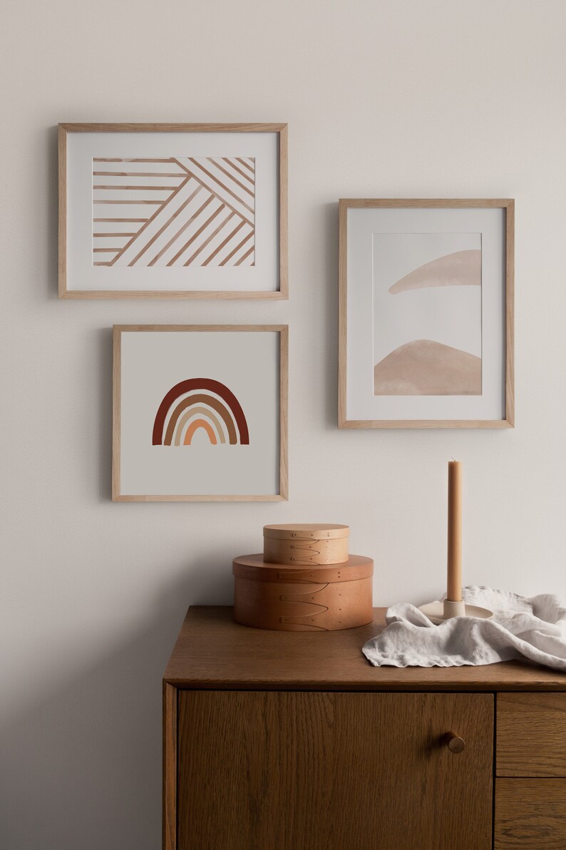 May include: Three framed abstract art prints with a minimalist design. The prints feature geometric shapes and lines in shades of brown and beige. The prints are displayed on a white wall above a wooden dresser.