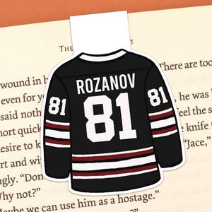 May include: A black and white hockey jersey bookmark with the name "ROZANOV" and the number "81". The bookmark is placed on an open book with text visible. The jersey has burgundy stripes.