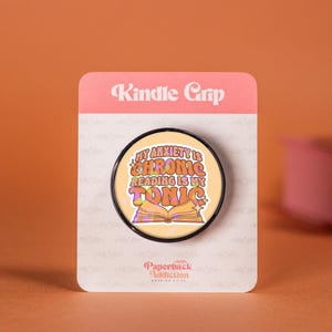 Puede incluir: Un agarre para Kindle redondo con el texto "My Anxiety is Chronic Reading is My Tonic" en naranja y amarillo. El agarre está en una tarjeta blanca con las palabras "Kindle Grip" en la parte superior. El fondo es naranja.